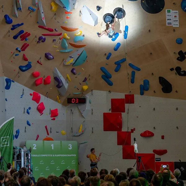 Een indoor klimwedstrijd met een klimmer op een kleurrijke klimwand, een menigte die beneden toekijkt en een podium dat aan de zijkant te zien is. Felle lampen belichten de klimmer en de klimgrepen.
